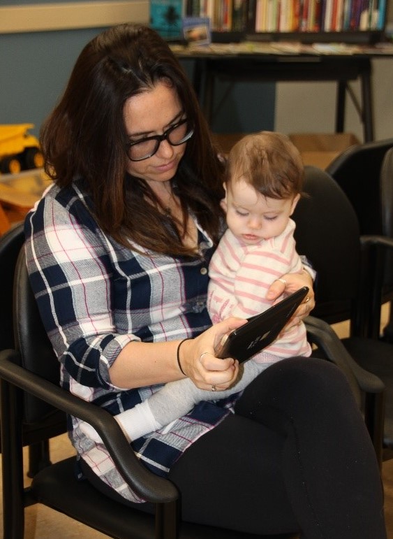 A mother holds her baby on her lap in a waiting room. Mother and baby are looking at a tablet, which the mother is holding.