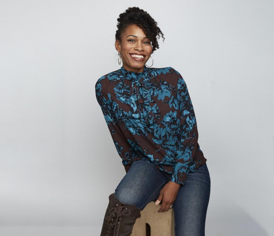 Bee is smiling. She has an updo and is wearing hoop earrings, a blue and brown floral top with jeans and brown boots. She sits on a stool at an angle.