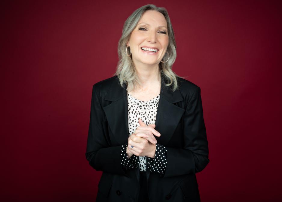 Nina smiles with her hands clasped in front. She stands in front of a dark red background and is wearing a black jacket with white spotted top.