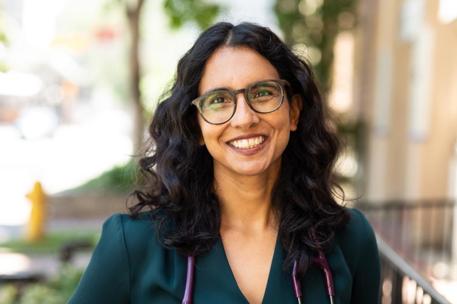Tara Kiran smiles at the camera. She is wearing glasses and her curly hair is in a centre part. She has on a dark green top, purple stethoscope and is standing outside, blurred background.