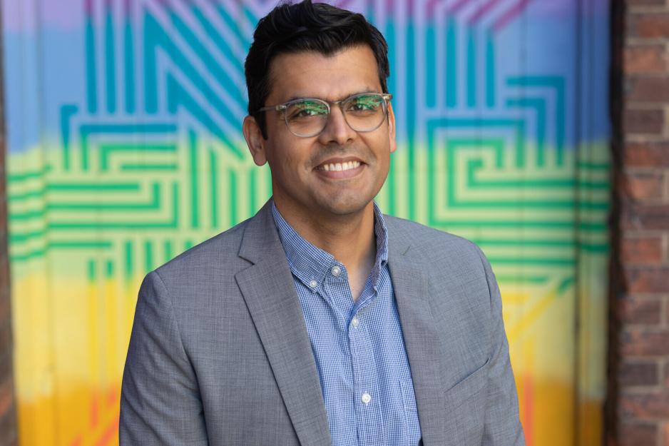 Danyaal Raza smiling in glasses and a grey suit in front a rainbow-coloured background.