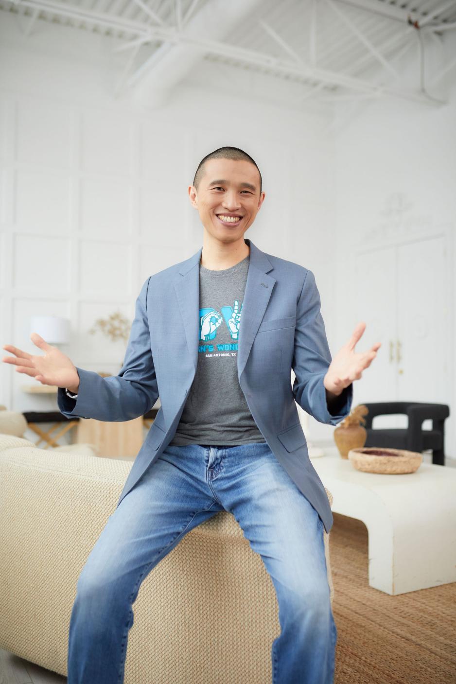 Terrence sits on the corner of a couch in jeans and jacket, arms spread, smiling